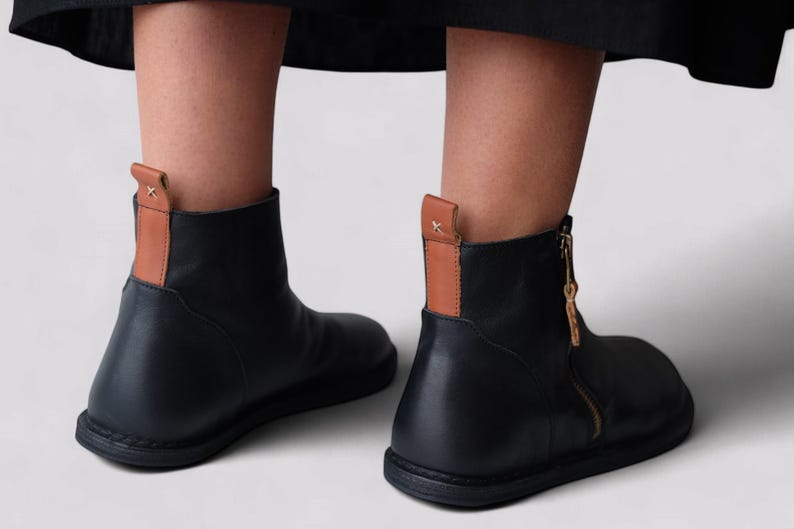 Pu&ograve; includere: Stivali da caviglia in pelle nera con un accento in pelle marrone sul retro e una cerniera laterale.