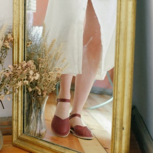 Pu&ograve; includere: Il riflesso di una donna in uno specchio con cornice dorata, che indossa un abito bianco e sandali in pelle bordeaux con cinturini alla caviglia.