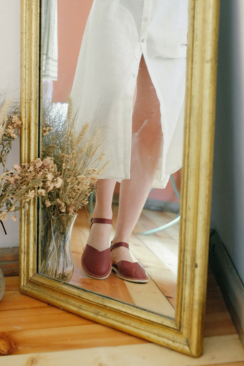 Pu&ograve; includere: Il riflesso di una donna in uno specchio con cornice dorata, che indossa un abito bianco e sandali in pelle bordeaux con cinturini alla caviglia.