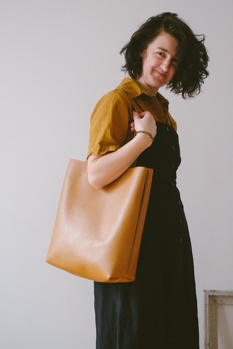 May include: A woman wearing a mustard yellow shirt and a black dress is holding a brown leather tote bag.