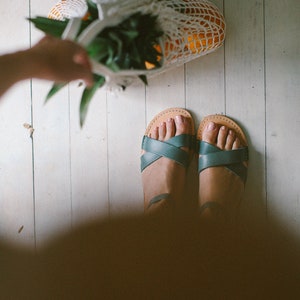 May include: A pair of teal leather sandals with criss-cross straps on a white wood floor. A mesh bag filled with oranges and a pineapple is in the background.