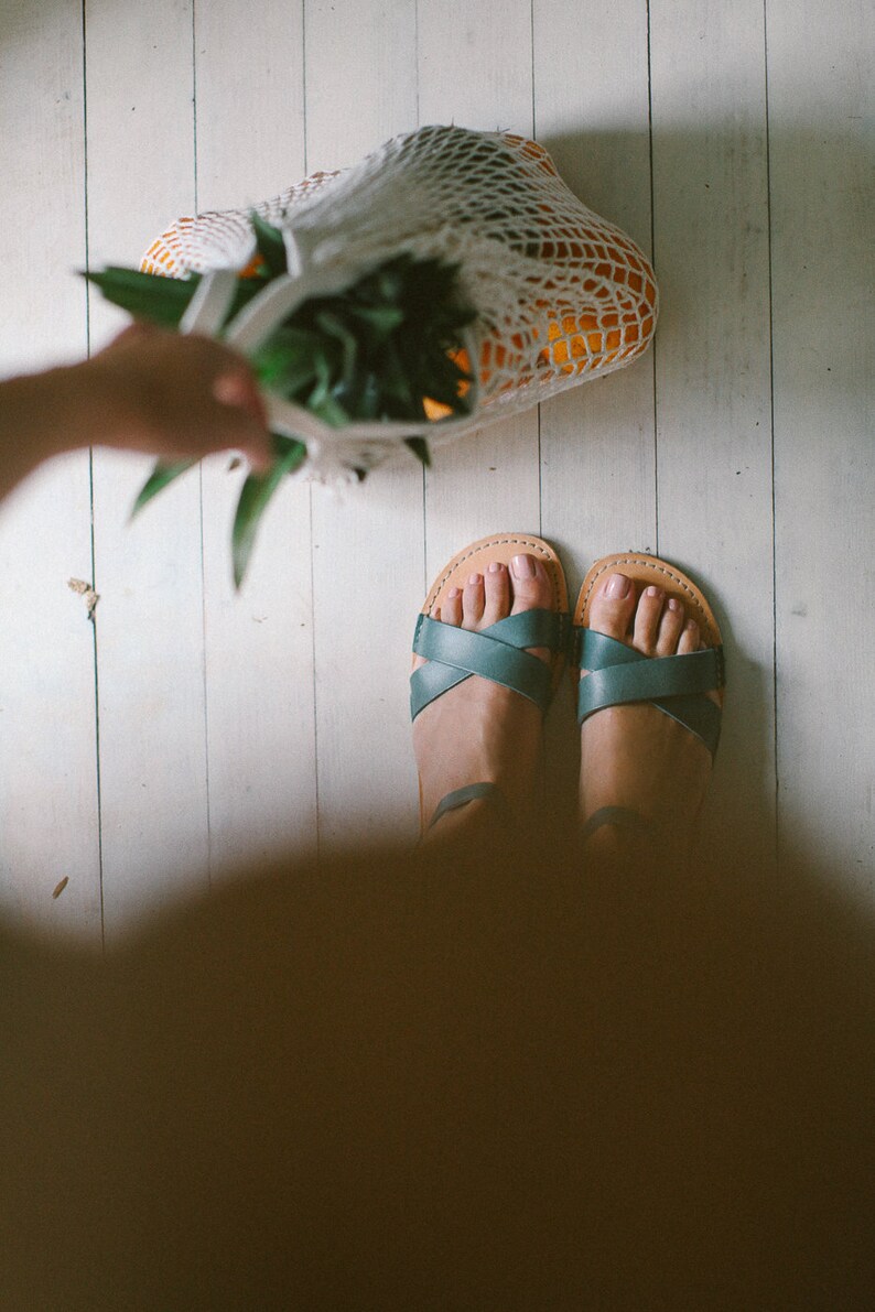 May include: A pair of teal leather sandals with criss-cross straps on a white wood floor. A mesh bag filled with oranges and a pineapple is in the background.