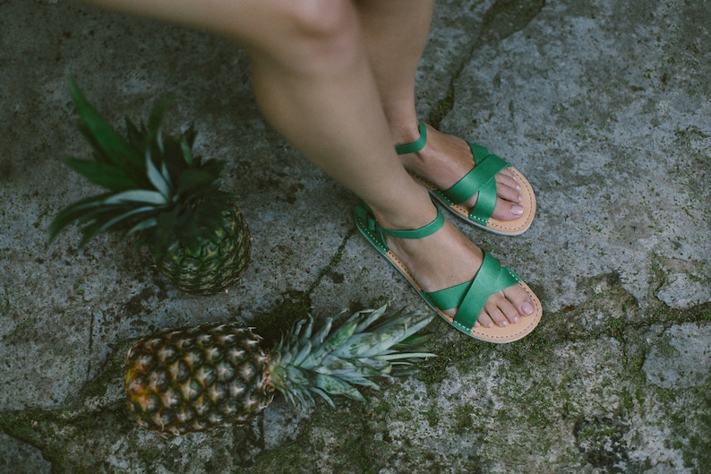 May include: A pair of green leather sandals with criss-cross straps. The sandals are on a concrete surface with two pineapples.
