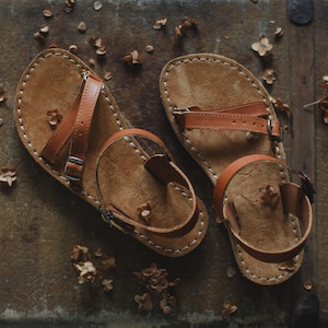 Peut inclure: Une paire de sandales en cuir faites &agrave; la main avec des lani&egrave;res et des semelles marron. Les sandales ont un design simple avec une boucle d'orteil et une bride de cheville, et sont cousues avec du fil blanc. Les sandales sont pr&eacute;sent&eacute;es sur une surface en bois.