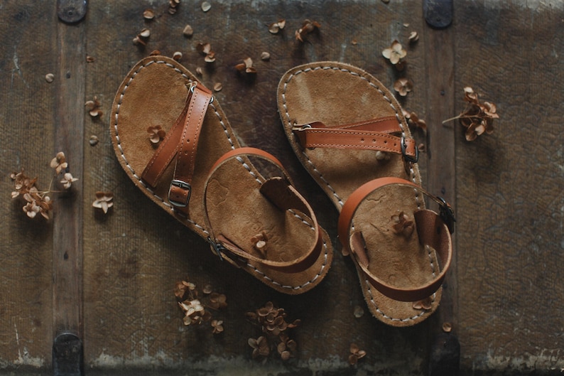 Peut inclure: Une paire de sandales en cuir faites &agrave; la main avec des lani&egrave;res et des semelles marron. Les sandales ont un design simple avec une boucle d'orteil et une bride de cheville, et sont cousues avec du fil blanc. Les sandales sont pr&eacute;sent&eacute;es sur une surface en bois.