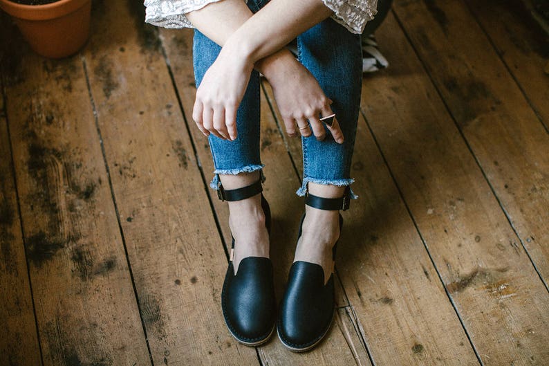 May include: A pair of black leather ankle strap sandals with a closed toe. The sandals are worn by a person sitting on a wooden floor.