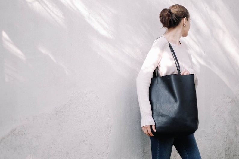 May include: A woman wearing a light pink sweater and blue jeans is holding a black leather tote bag.