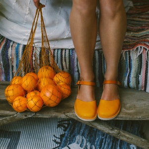 May include: A woman wearing a floral dress and yellow leather sandals holds a mesh bag filled with oranges. The bag is sitting on a wooden surface with a striped rug underneath.
