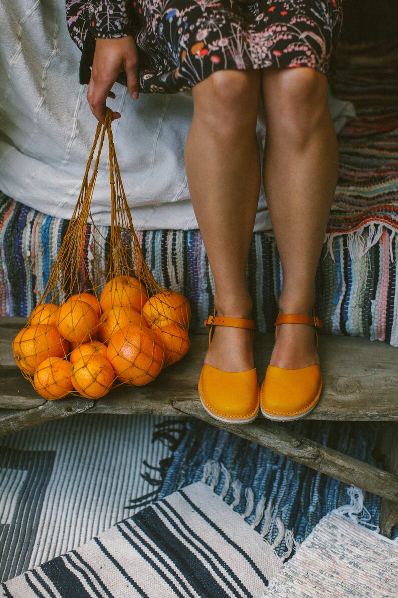 May include: A woman wearing a floral dress and yellow leather sandals holds a mesh bag filled with oranges. The bag is sitting on a wooden surface with a striped rug underneath.
