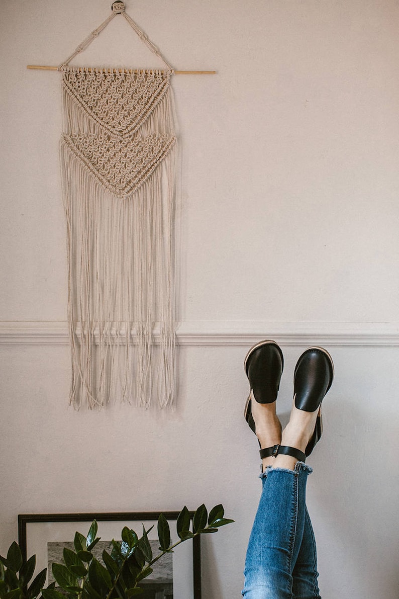 May include: A white macrame wall hanging with a geometric pattern hangs on a white wall. A pair of black leather ankle strap sandals with a wooden heel are propped up against the wall with the toes pointing up. The sandals are worn by a person wearing blue jeans.