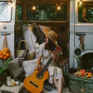 May include: A woman wearing a straw hat sits in a vintage van playing an acoustic guitar. The van is decorated with string lights and pillows. There are fruits and a pineapple on a wooden crate in the foreground.