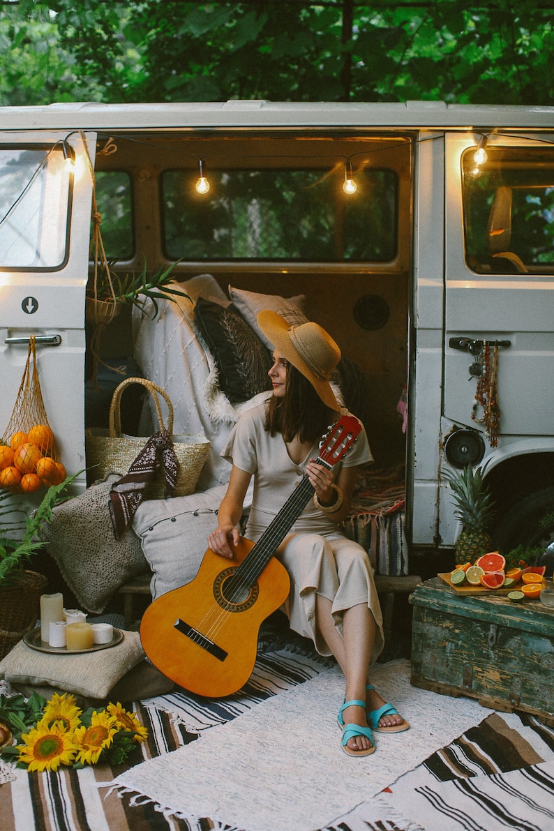 May include: A woman wearing a straw hat sits in a vintage van playing an acoustic guitar. The van is decorated with string lights and pillows. There are fruits and a pineapple on a wooden crate in the foreground.