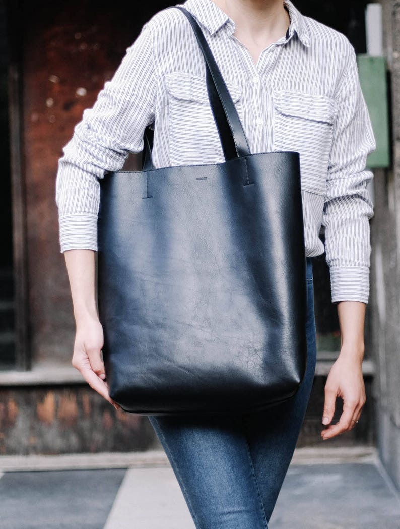 May include: A person wearing a striped button-down shirt and blue jeans is carrying a large black leather tote bag with a long strap.