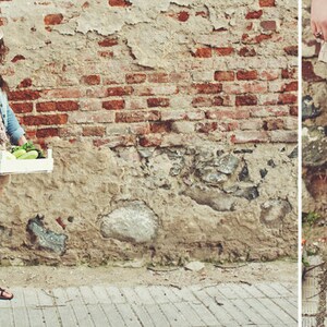 May include: A person wearing a denim jacket, shorts, and sandals carries a white wooden crate of vegetables and a crocheted tote bag. The tote bag is filled with bread. The background is a weathered brick wall.