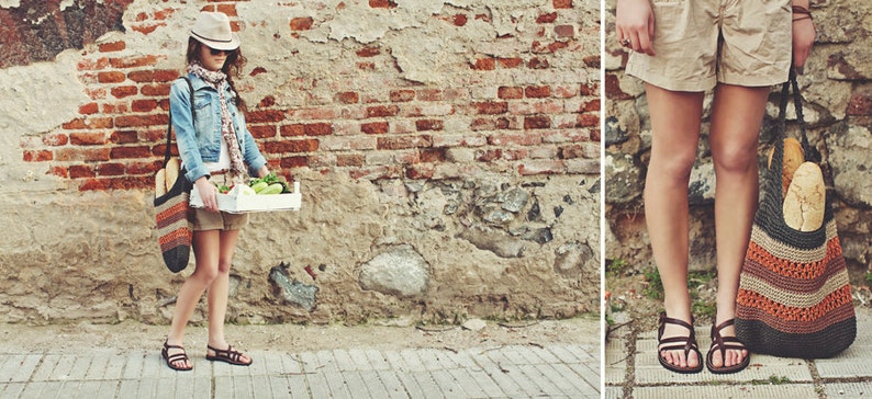 May include: A person wearing a denim jacket, shorts, and sandals carries a white wooden crate of vegetables and a crocheted tote bag. The tote bag is filled with bread. The background is a weathered brick wall.