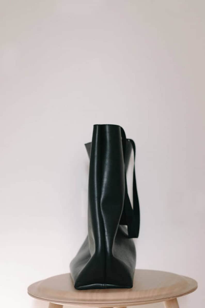 May include: A black leather tote bag with a single strap. The bag is sitting on a light wood table.