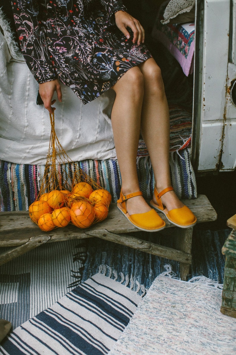 May include: A woman sits on a bed with a mesh bag filled with oranges. She is wearing a floral dress and yellow sandals. The bed is covered in a white blanket and a colorful striped rug.