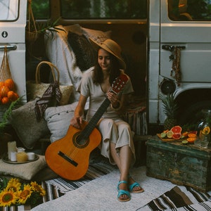 May include: A woman sits in front of a vintage van, playing an acoustic guitar. She is wearing a straw hat and a beige dress. The van is decorated with string lights and a variety of plants and fruit.