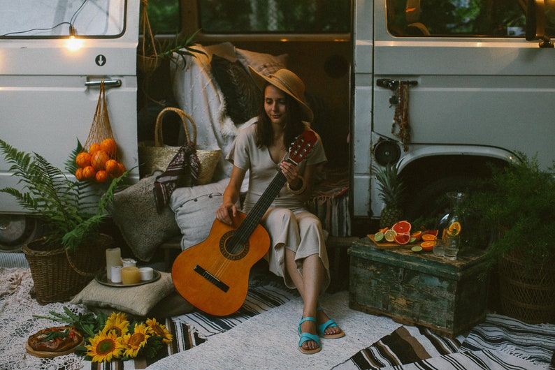 May include: A woman sits in front of a vintage van, playing an acoustic guitar. She is wearing a straw hat and a beige dress. The van is decorated with string lights and a variety of plants and fruit.