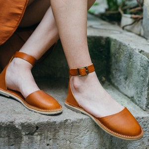 May include: A pair of brown leather sandals with ankle straps and buckles. The sandals have a flat sole and are made of a soft, supple leather.