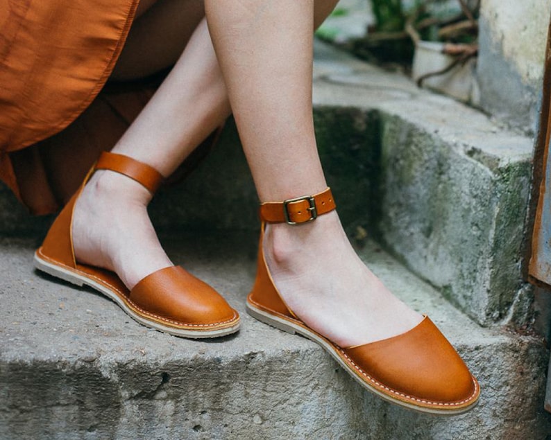May include: A pair of brown leather sandals with ankle straps and buckles. The sandals have a flat sole and are made of a soft, supple leather.
