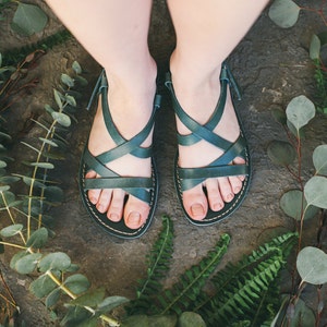 May include: A pair of dark green leather sandals with criss-cross straps. The sandals are on a stone surface surrounded by green foliage.