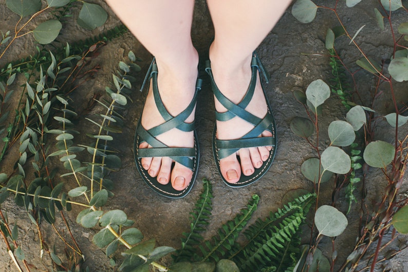 May include: A pair of dark green leather sandals with criss-cross straps. The sandals are on a stone surface surrounded by green foliage.