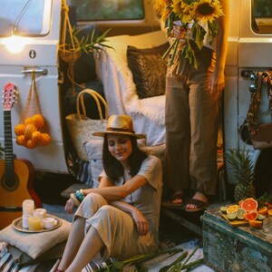 May include: Two women in a vintage van, one wearing a straw hat and holding a bunch of sunflowers, the other sitting on a blanket with a bunch of sunflowers at her feet.
