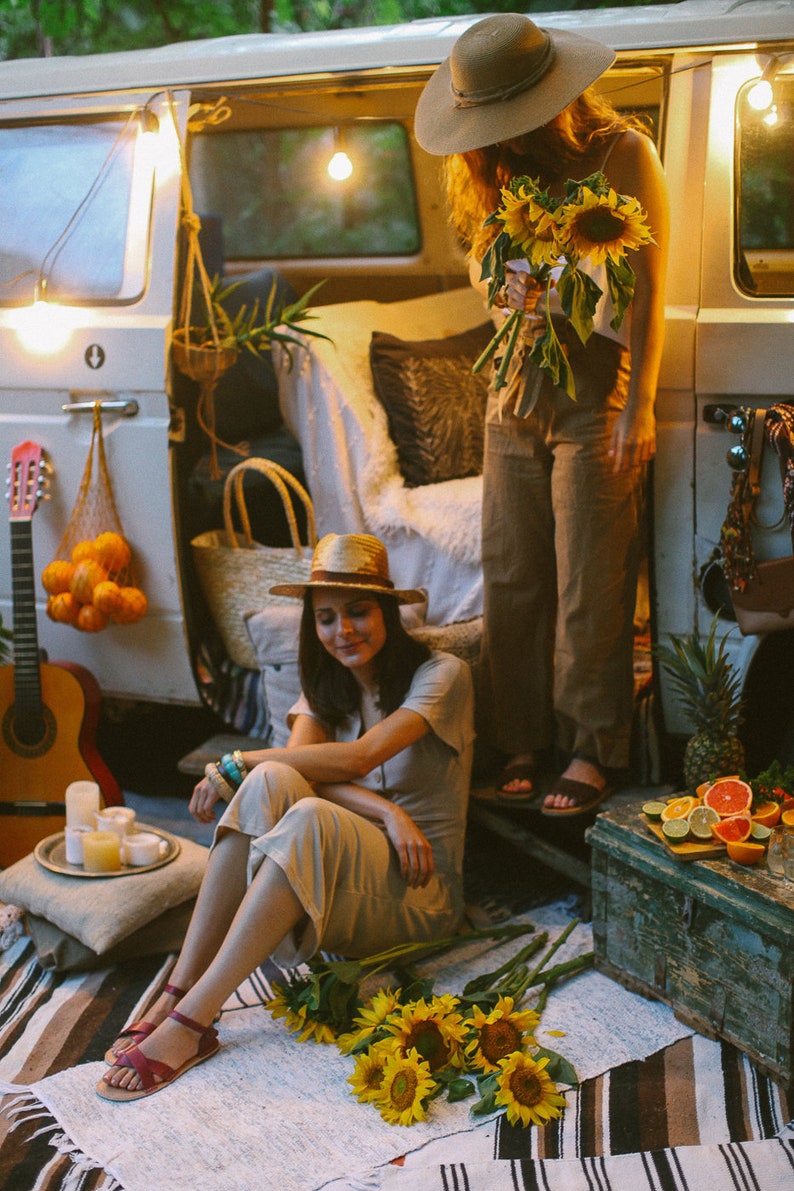 May include: Two women in a vintage van, one wearing a straw hat and holding a bunch of sunflowers, the other sitting on a blanket with a bunch of sunflowers at her feet.