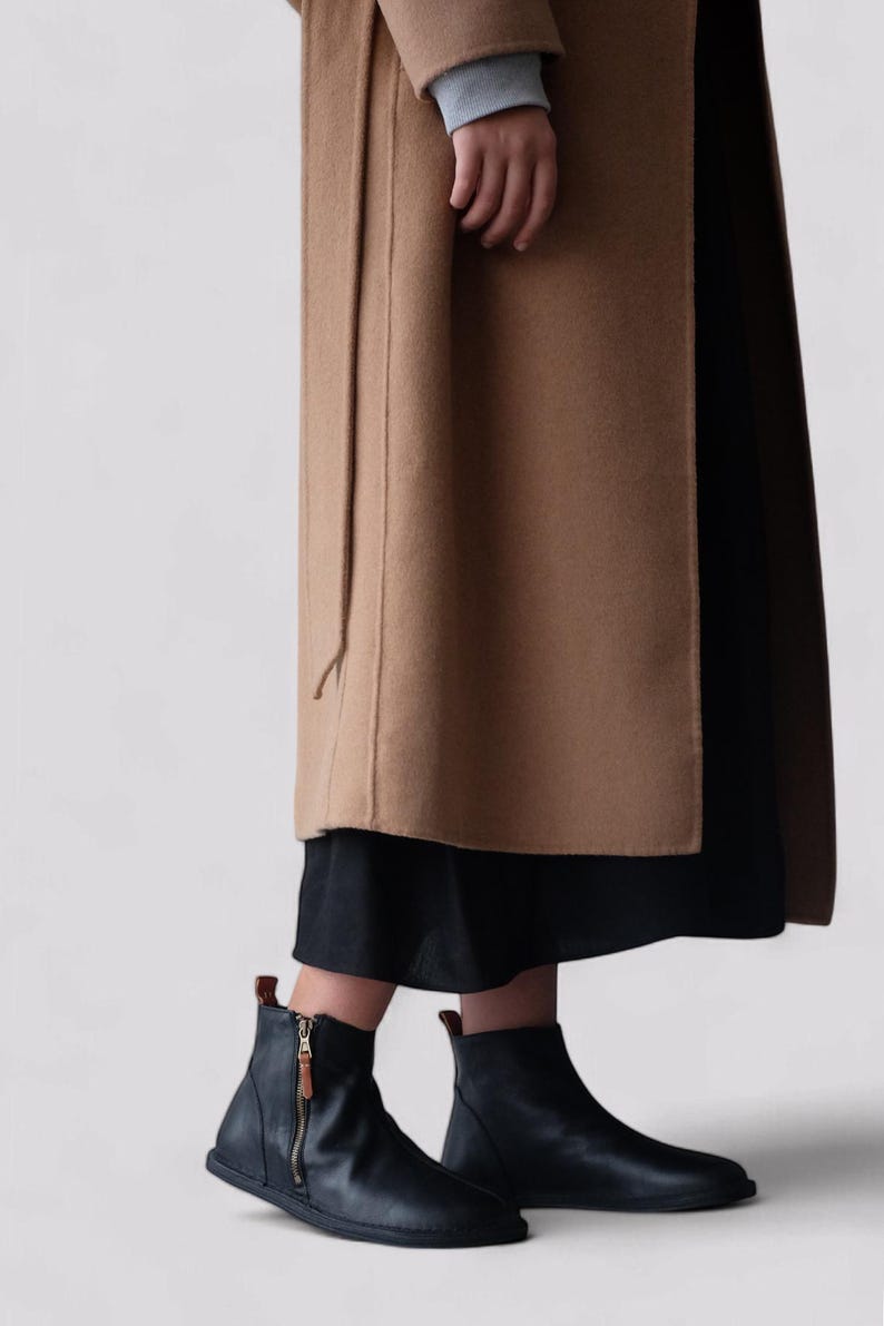 Pu&ograve; includere: Stivali neri in pelle con cerniera laterale e punta arrotondata. Gli stivali sono indossati con un cappotto marrone e una gonna nera.