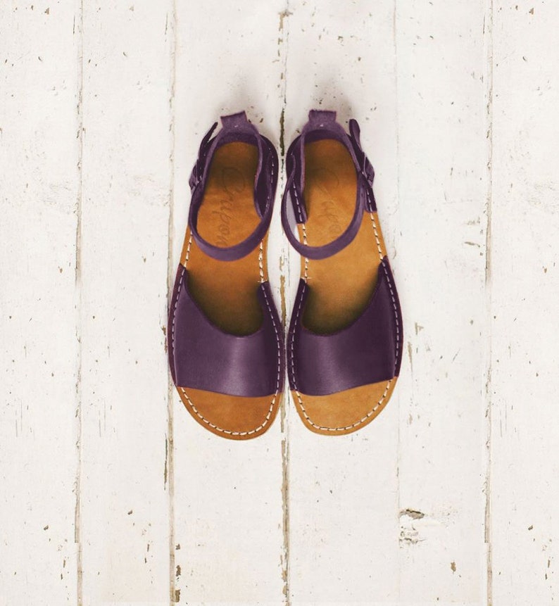 May include: A pair of purple leather sandals with ankle straps. The sandals have a brown leather sole and are photographed against a white wooden backdrop.