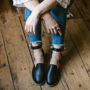 Op de afbeelding: Een paar zwarte leren enkelband sandalen met een gesloten neus. De sandalen worden gedragen door een persoon die op een houten vloer zit.