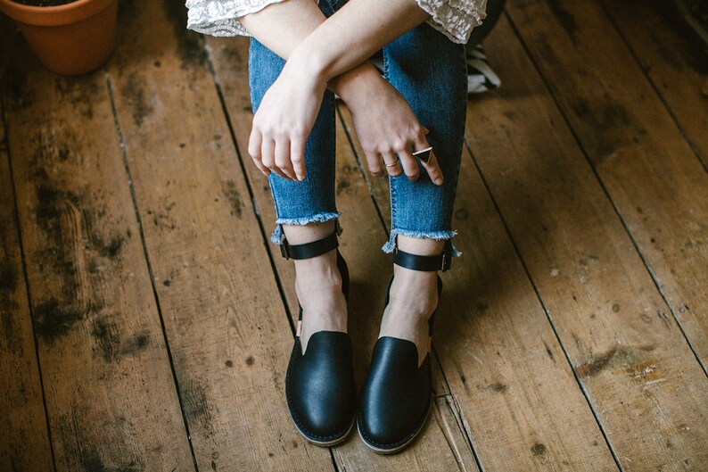 Op de afbeelding: Een paar zwarte leren enkelband sandalen met een gesloten neus. De sandalen worden gedragen door een persoon die op een houten vloer zit.
