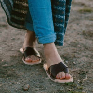 May include: A pair of black leather sandals with a criss-cross strap design. The sandals are worn on a person's feet, with the person's blue jeans and a patterned skirt visible.