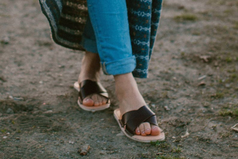 May include: A pair of black leather sandals with a criss-cross strap design. The sandals are worn on a person's feet, with the person's blue jeans and a patterned skirt visible.