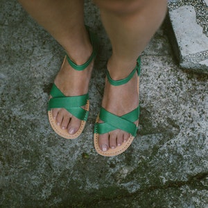 May include: A pair of green leather sandals with criss-cross straps. The sandals are worn on a person's feet.