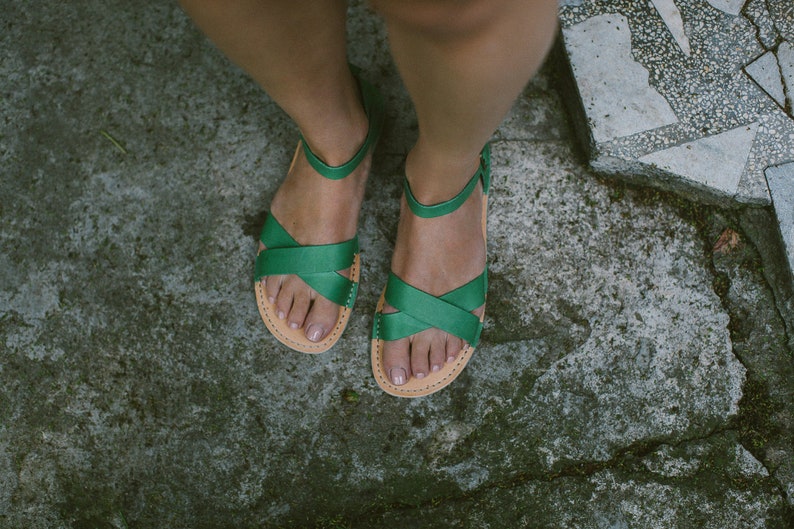 May include: A pair of green leather sandals with criss-cross straps. The sandals are worn on a person's feet.