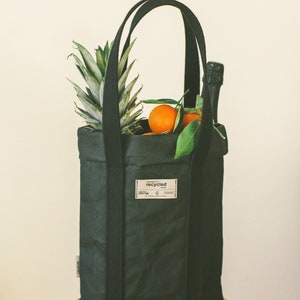 May include: A black reusable tote bag with thick straps, filled with a pineapple, oranges, and a bottle of champagne. The bag has a label that reads "HANDMADE OF recycled PAPER".