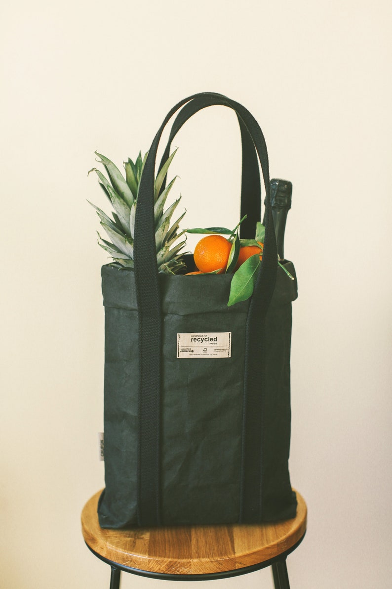 May include: A black reusable tote bag with thick straps, filled with a pineapple, oranges, and a bottle of champagne. The bag has a label that reads "HANDMADE OF recycled PAPER".
