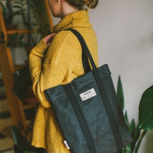 May include: A person wearing a yellow sweater and blue jeans is carrying a black tote bag with black straps. The bag has a white label that says "recycled".