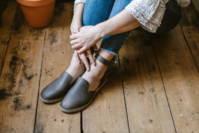 Op de afbeelding: Een paar grijze leren enkelband sandalen met een simpel ontwerp. De sandalen worden gedragen door een persoon die op een houten vloer zit.