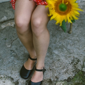 Pode incluir: Uma mulher vestindo um vestido de bolinhas vermelhas e brancas e sand&aacute;lias de couro pretas senta-se em uma cadeira segurando um girassol.