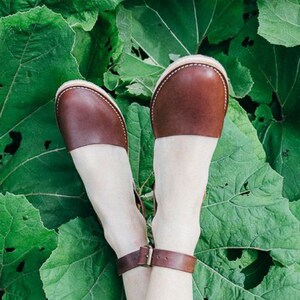 May include: A pair of brown leather sandals with ankle straps. The sandals are on a green leafy background.