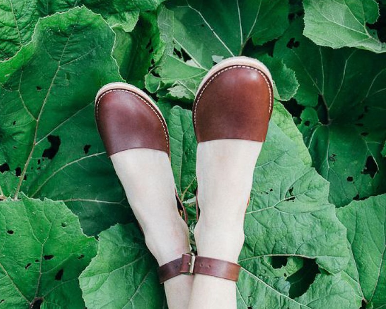 May include: A pair of brown leather sandals with ankle straps. The sandals are on a green leafy background.