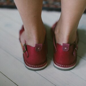 May include: A pair of burgundy leather sandals with a buckle fastening. The sandals have a white stitching detail around the sole.