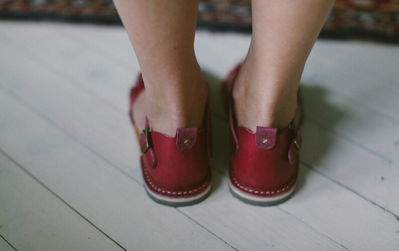May include: A pair of burgundy leather sandals with a buckle fastening. The sandals have a white stitching detail around the sole.