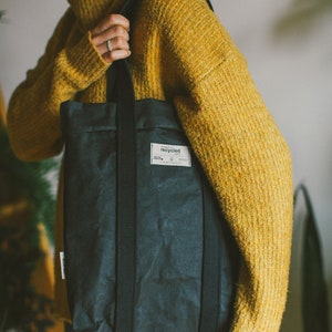 May include: A person wearing a yellow sweater is holding a black tote bag with a label that says "recycled".