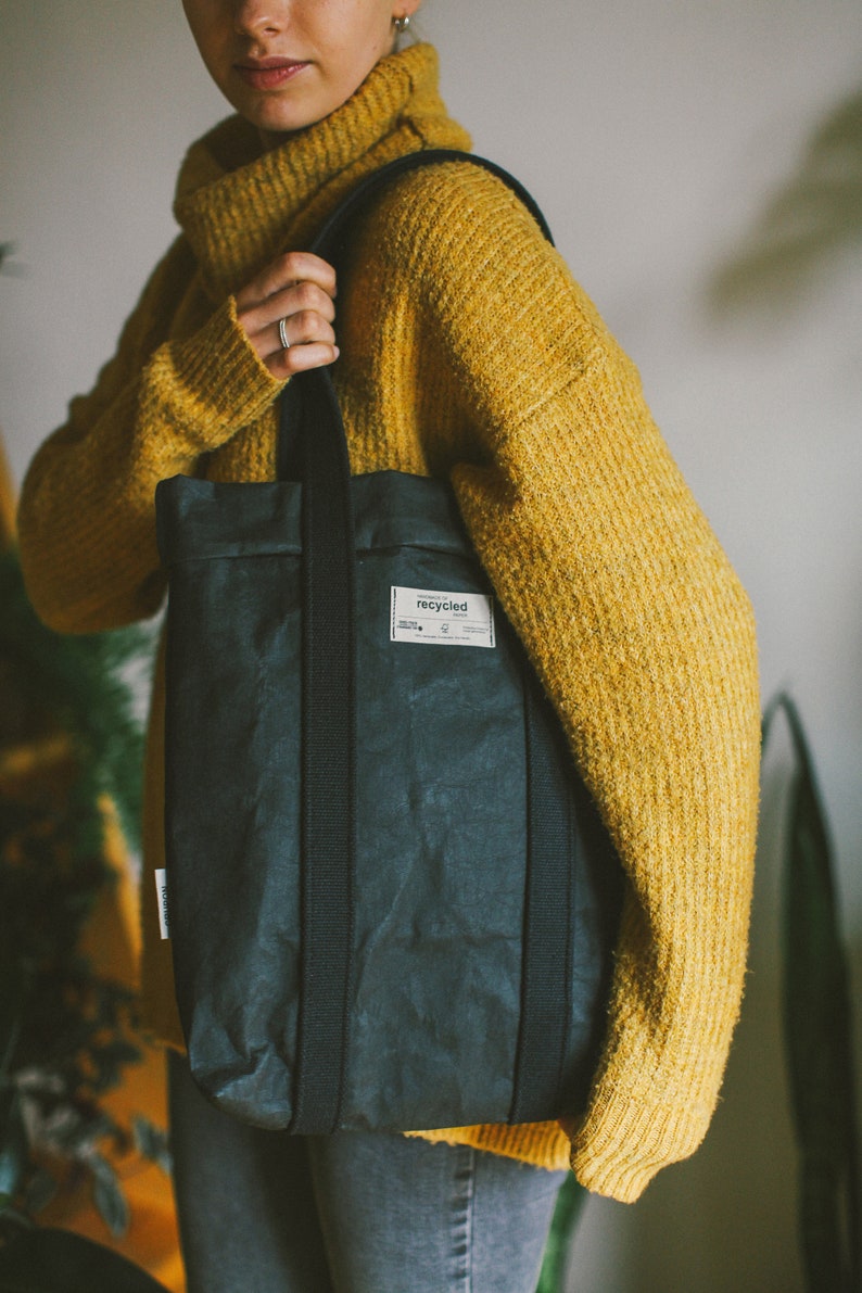 May include: A person wearing a yellow sweater is holding a black tote bag with a label that says "recycled".
