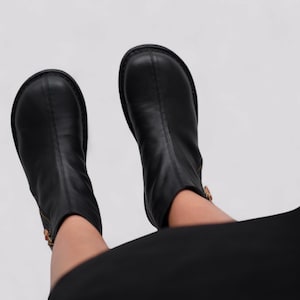 Pu&ograve; includere: Un paio di stivaletti neri in pelle con un dettaglio di cerniera laterale. Gli stivaletti sono indossati da una persona che indossa pantaloni neri.