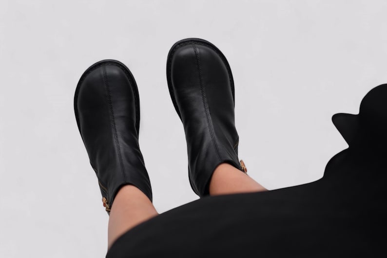 Pu&ograve; includere: Un paio di stivaletti neri in pelle con un dettaglio di cerniera laterale. Gli stivaletti sono indossati da una persona che indossa pantaloni neri.